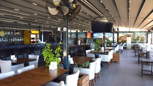 A restaurant with multiple tables and chairs, a bar area with bottles, and a large fan. - Sultans And Kings Hotel (Didim)