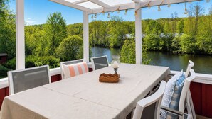 Un espacio cubierto para comer al aire libre con una mesa, sillas y vistas a un río y un bosque.