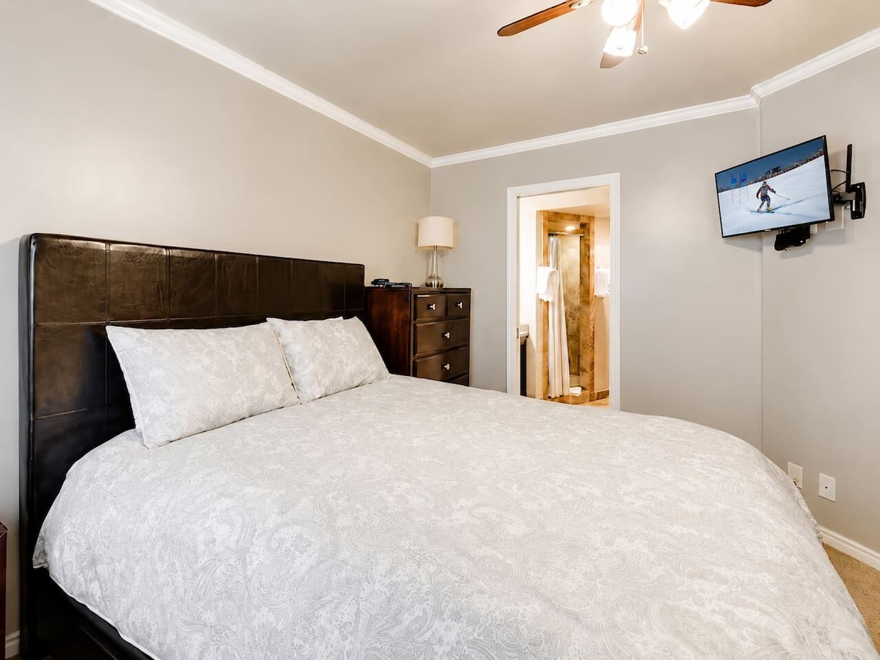 A bedroom with a large bed, a dresser, a TV mounted on the wall, and a door leading to a bathroom.