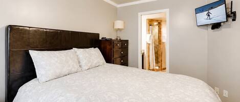 A bedroom with a large bed, a dresser, a TV mounted on the wall, and a door leading to a bathroom.