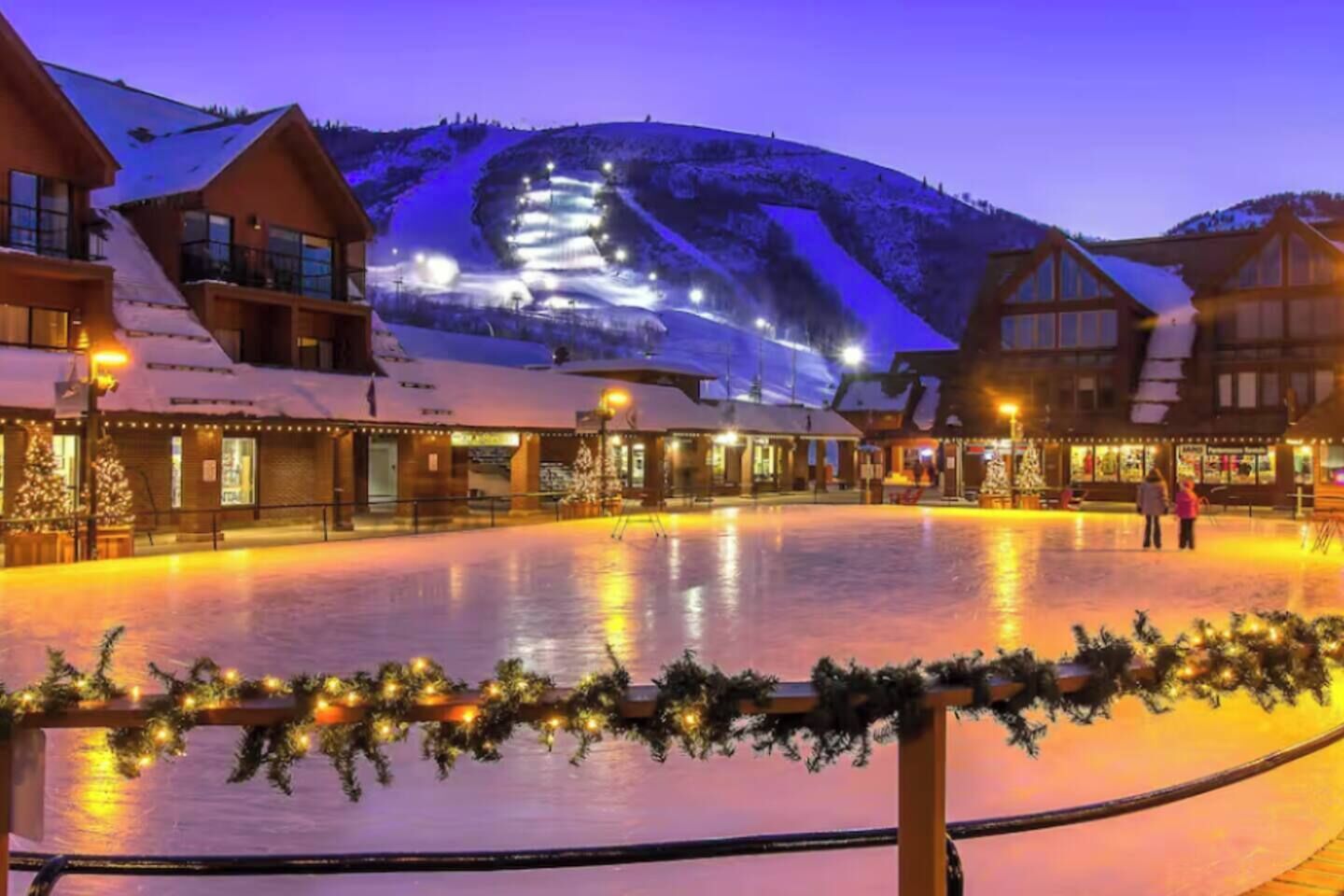 A snow-covered outdoor area with buildings, a ski slope in the background, and a Christmas tree.