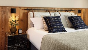 Quarto com cabeceira de madeira, mesa de cabeceira de mármore, cama com roupa de cama branca e almofadas estampadas, um vaso com flores e um tapete texturizado.