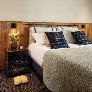 Quarto com cabeceira de madeira, mesa de cabeceira de mármore, cama com roupa de cama branca e almofadas estampadas, um vaso com flores e um tapete texturizado.