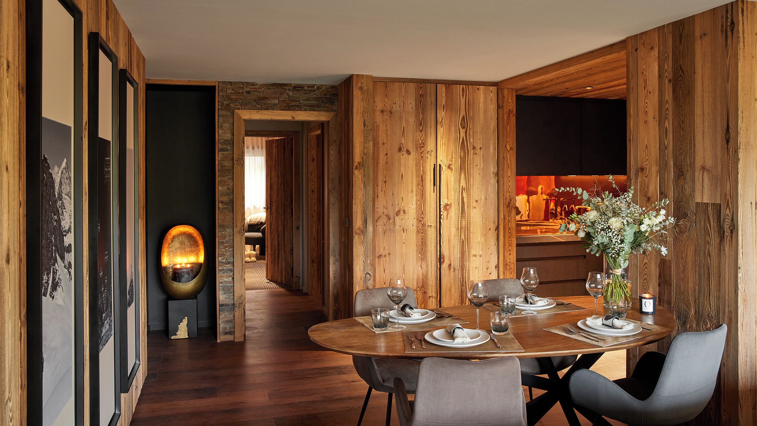 A dining area with a wooden table set for four, a vase with flowers, and a fireplace in the background.
