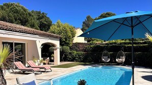 Une piscine entourée d’un jardin et d’un bâtiment, avec des chaises longues et un parasol à proximité.