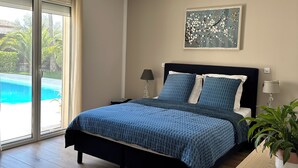 Une chambre à coucher comprenant un lit, un bureau, une chaise, une lampe, une plante et une fenêtre donnant sur une piscine.