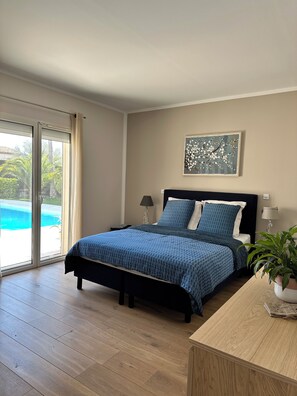 Ein Schlafzimmer mit Bett, Schreibtisch, Stuhl, Lampe, Pflanze und einem Fenster mit Blick auf einen Pool.