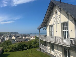A two-story building with a balcony, a clear view of the sea, and a town in the background. - Normandy seaside villa with view and terrace 5 minutes walk from the beach (Arromanches-les-Bains)