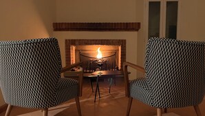 Une pièce avec une cheminée, deux fauteuils à motifs, une table et une fenêtre.