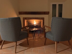 Ein Zimmer mit Kamin, zwei gemusterte Sessel, einem Tisch und einem Fenster.
