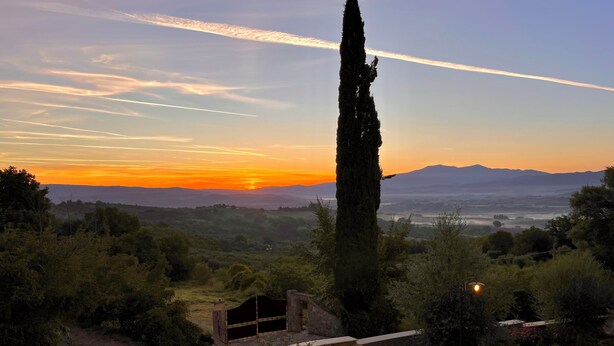 Un paysage comprenant un grand arbre, une structure en pierre et une vue sur des montagnes lointaines au coucher du soleil.