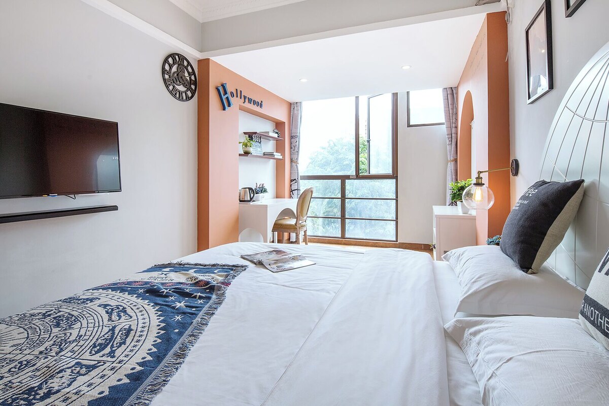 A bedroom with a bed, a TV, a clock, a window, and a desk area.
