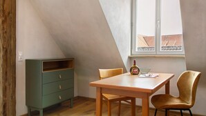 A room with a sloped ceiling, a wooden table with chairs, a green chest of drawers, and a window with a view of buildings.
