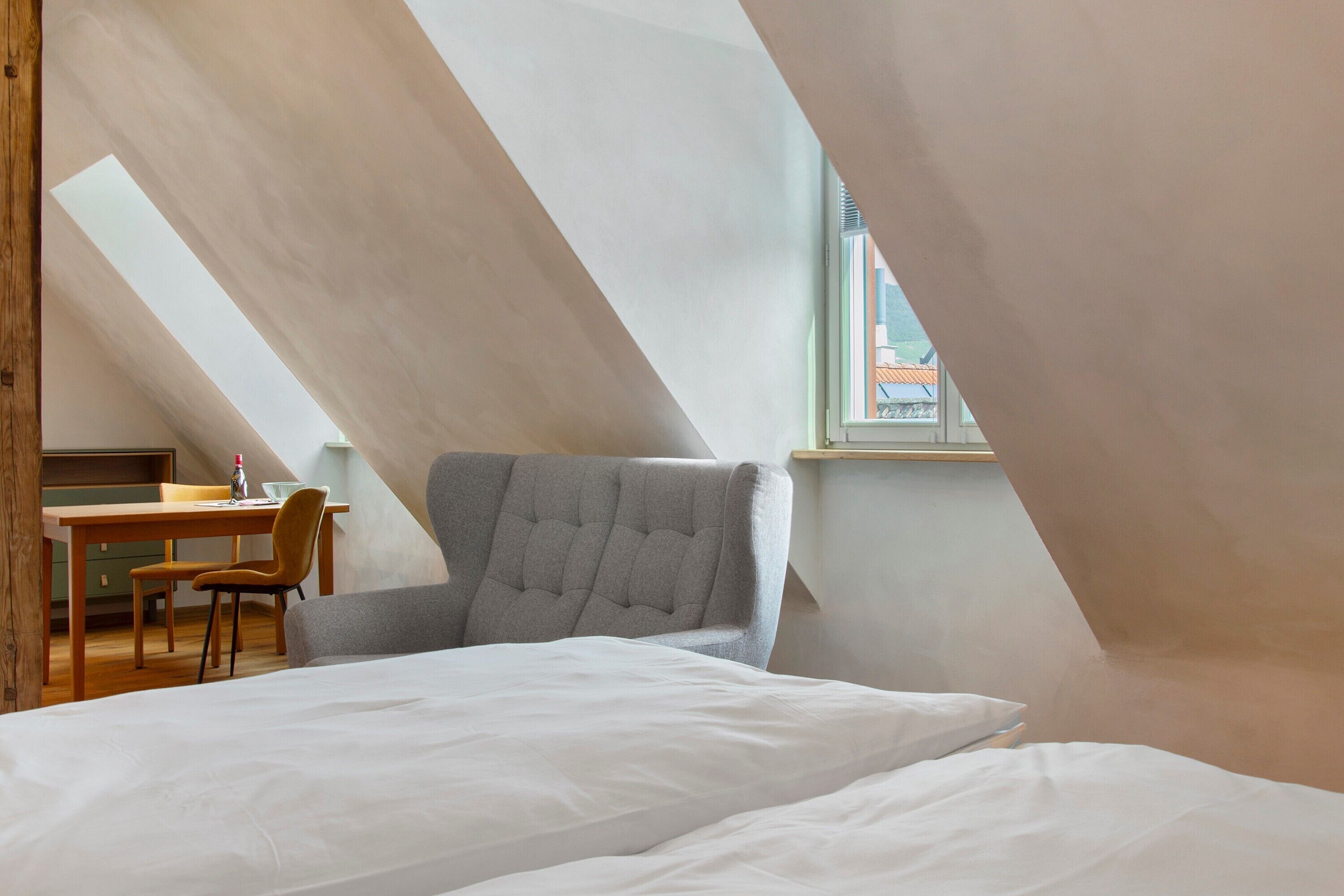 Un dormitorio con techo inclinado, una cama, una silla, un escritorio y una ventana.
