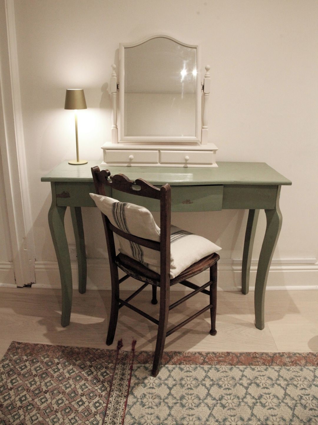 A room with a green desk, a chair with a cushion, a lamp, and a mirror.