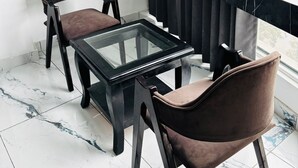 A balcony with a glass table and two chairs, a marble floor, and a view of a building. - Hotel The Amaresh (Panipat)