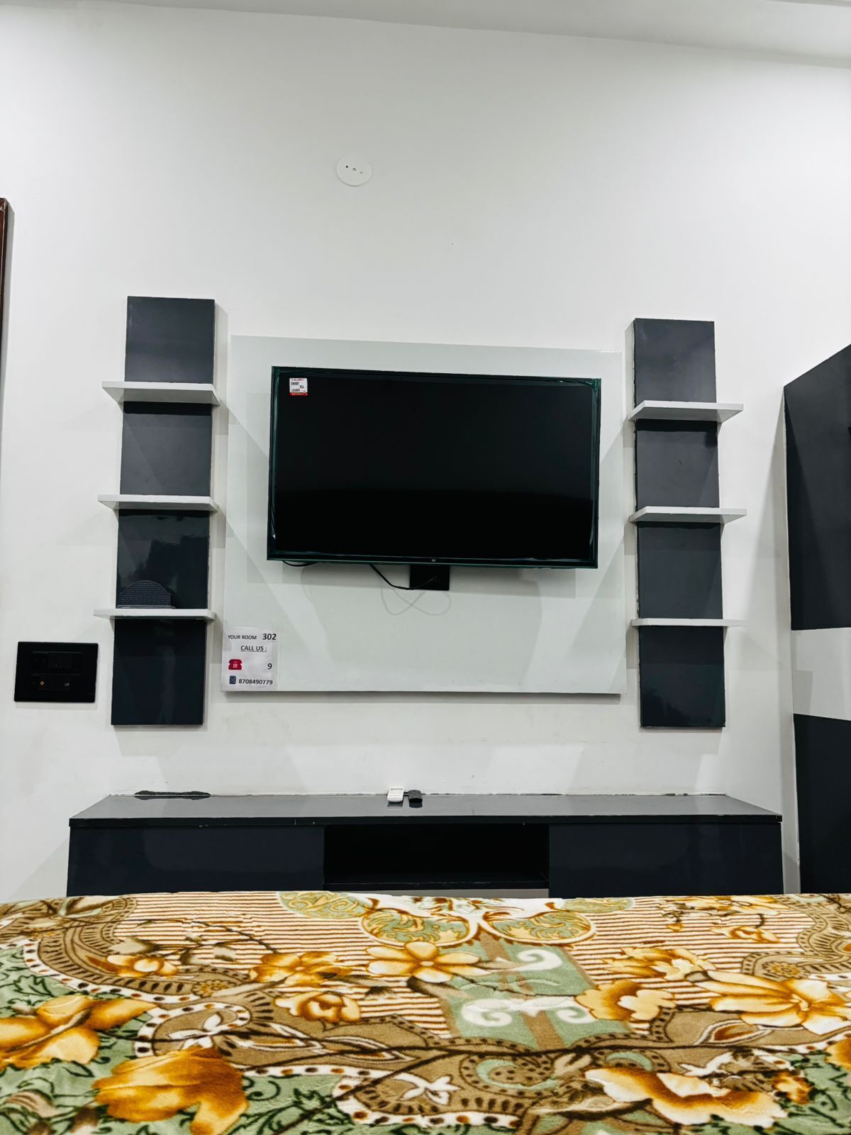 A room with a flat-screen TV mounted on the wall, a bed with a patterned bedspread, and a small shelf unit with a phone and a sign indicating room number 302.