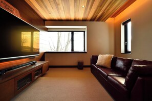 Ein modernes Wohnzimmer mit Flachbildfernseher, Holzbalkendecke, großen Fenstern, einer Ledersofa und einem kleinen Beistelltisch.