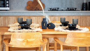 A wooden dining table with chairs, a vase with decorative branches, and a modern kitchen in the background. - Sundaze Villas - Cozy Stay in Canggu (Kerobokan)