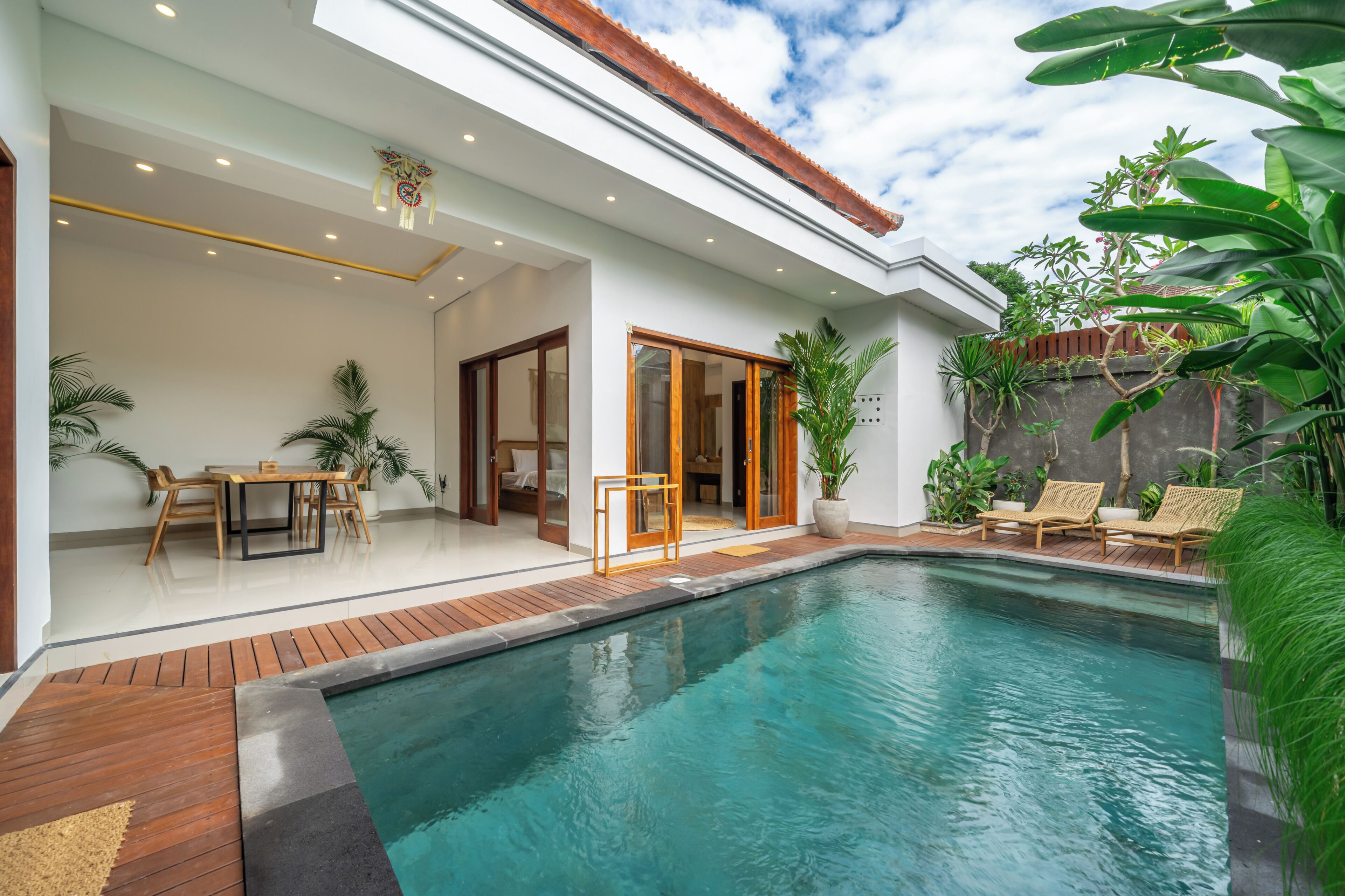 A swimming pool with wooden deck, a dining area with chairs and table, and a covered patio with a ceiling light.
