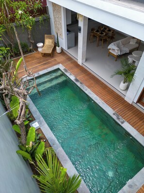 A rectangular swimming pool with a wooden deck, surrounded by greenery and a modern outdoor seating area with a sofa and chairs. - Sundaze Villas - Cozy Stay in Canggu (Kerobokan)