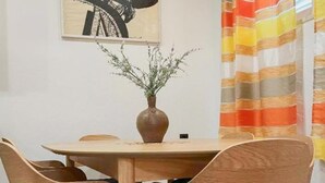 Een eetgedeelte met een ronde tafel, vier houten stoelen, een vaas met planten en een gestreept gordijn.