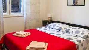 Een net opgemaakt bed met een rode sprei en stippilhoudende kussens, een nachtkastje met een lamp en een raam met gordijnen.