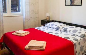 Ein ordentlich bezogenes Bett mit roter Tagesdecke und Kissen mit Punkten, ein Nachttisch mit Lampe und ein Fenster mit Vorhängen.