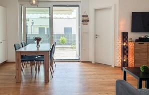 Ein Essbereich mit Holztisch und Stühlen, einer Vase mit Blumen und Blick durch eine Glastür auf einen Außenbereich mit einem Gebäude und einem Baum.