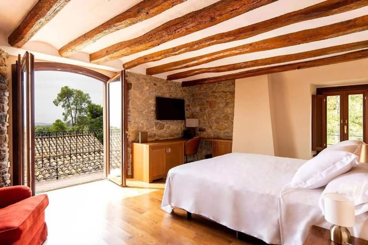 A bedroom with a stone wall, wooden beams, a bed, a desk, and a balcony with a view.