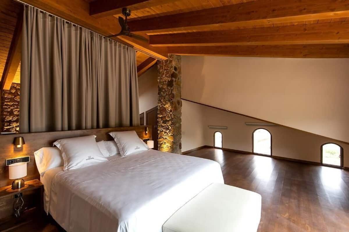 A bedroom with a large bed, wooden ceiling beams, stone wall, and skylight windows.