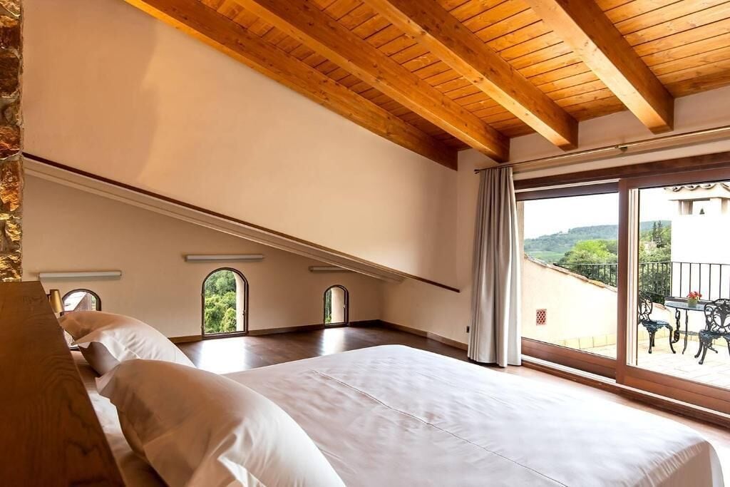A bedroom with a large bed, wooden ceiling beams, and a balcony with outdoor furniture.