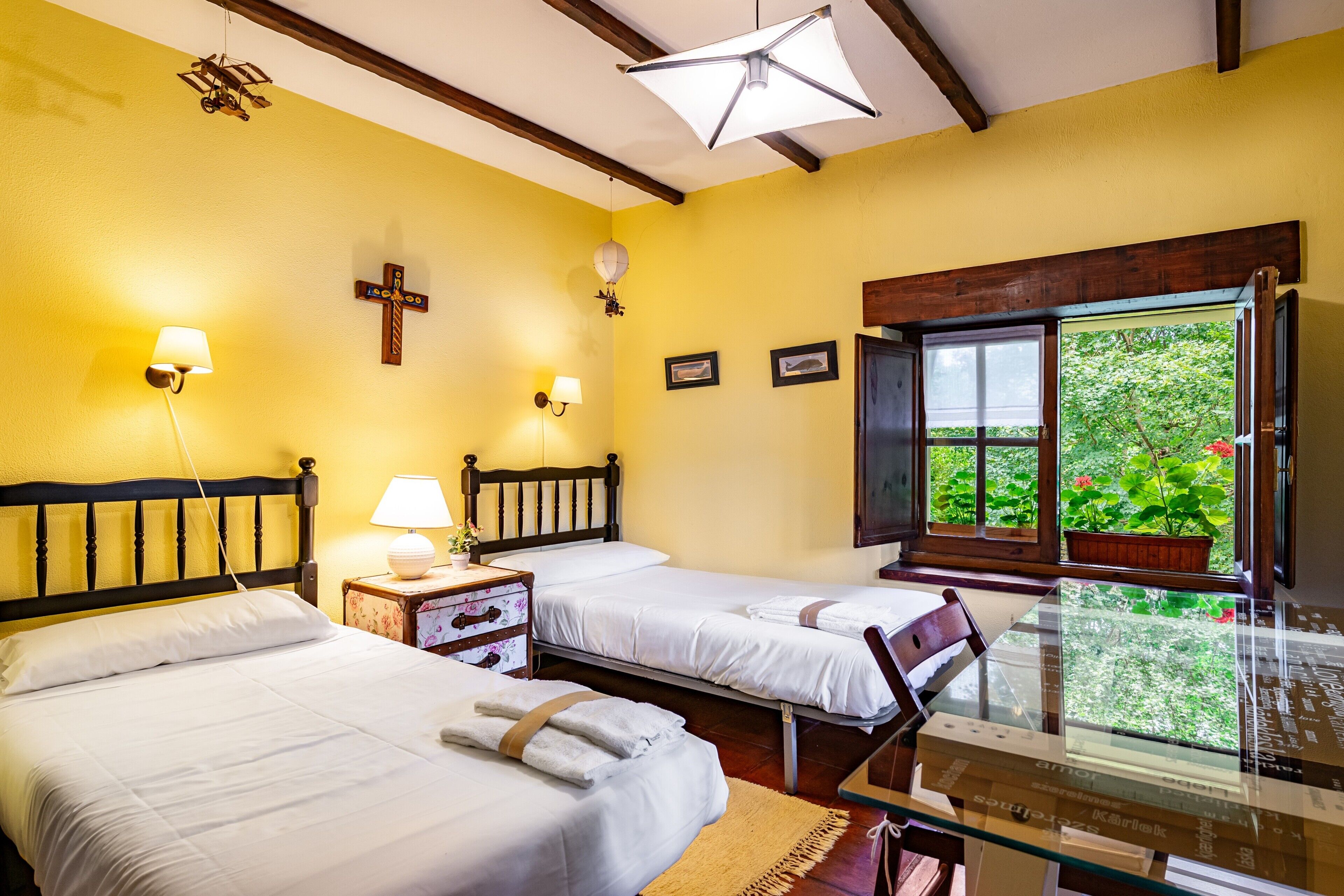 Une chambre avec deux lits, une tête de lit en bois, des lampes de chevet, une croix au mur et une fenêtre donnant sur un paysage verdoyant.