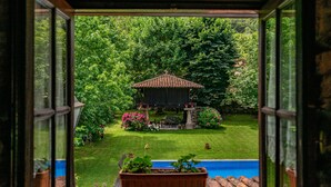Vista a través de una ventana que muestra un jardín con piscina, un pabellón y abundante vegetación.