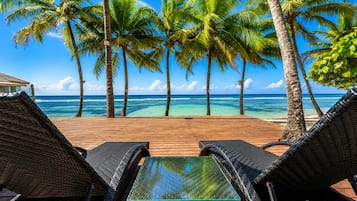 Een houten terras met rieten lounge fauteuils en een tafel met glazen blad, met uitzicht op een strand met palmbomen en helderblauw water.