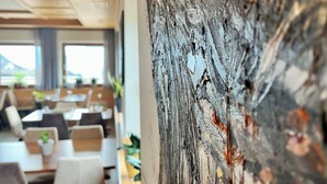 A dining area with tables and chairs, a large abstract painting on the wall, and a view of the outdoors through large windows. - Hotel Gasthof Kircher (Fie allo Sciliar)