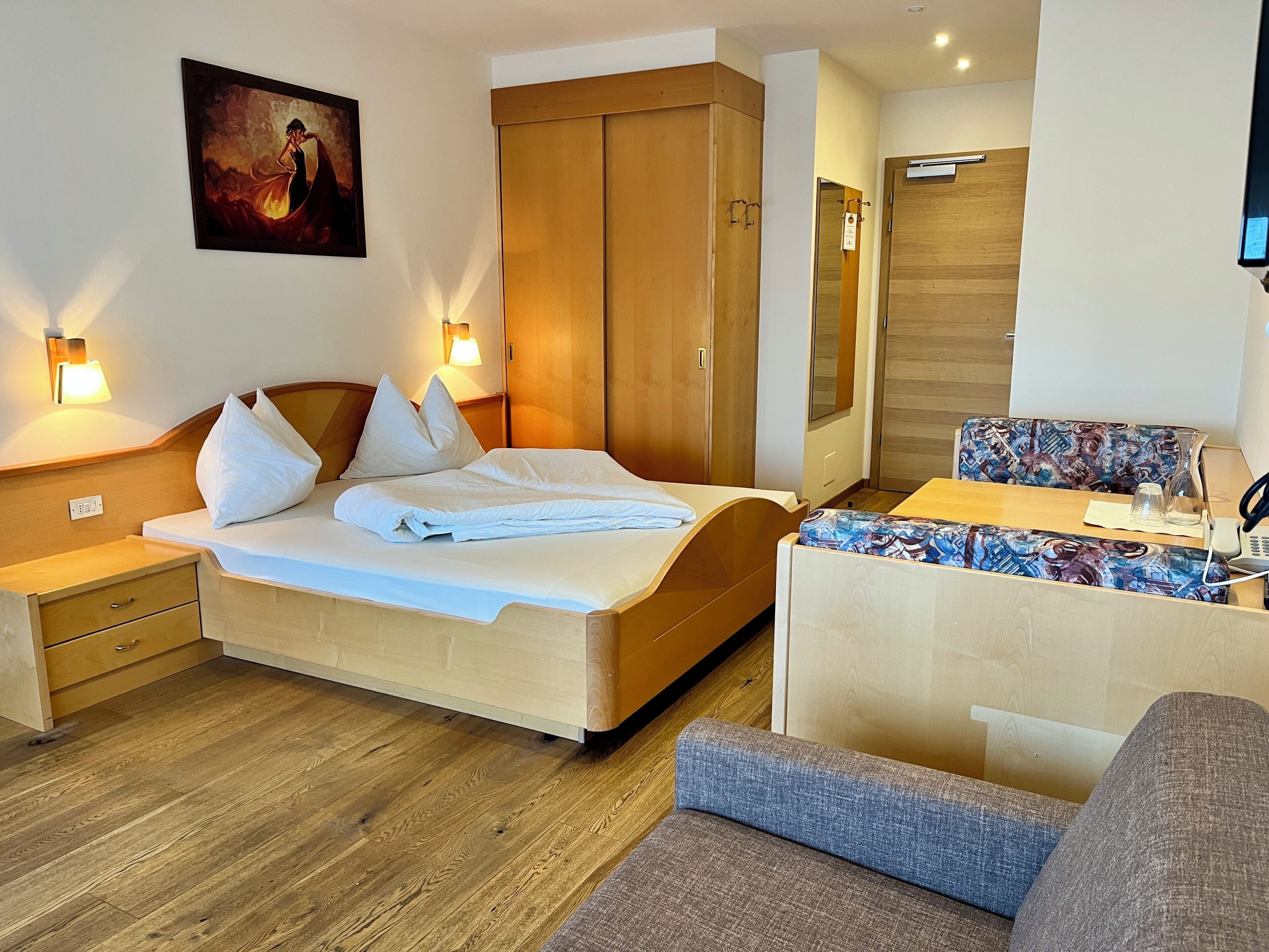 A hotel room with a bed, a wooden wardrobe, a desk with a chair, and a painting on the wall.