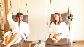 Two individuals in white robes on separate swings in a room with a wooden floor, a glass candle holder, and a vase with dried plants.
