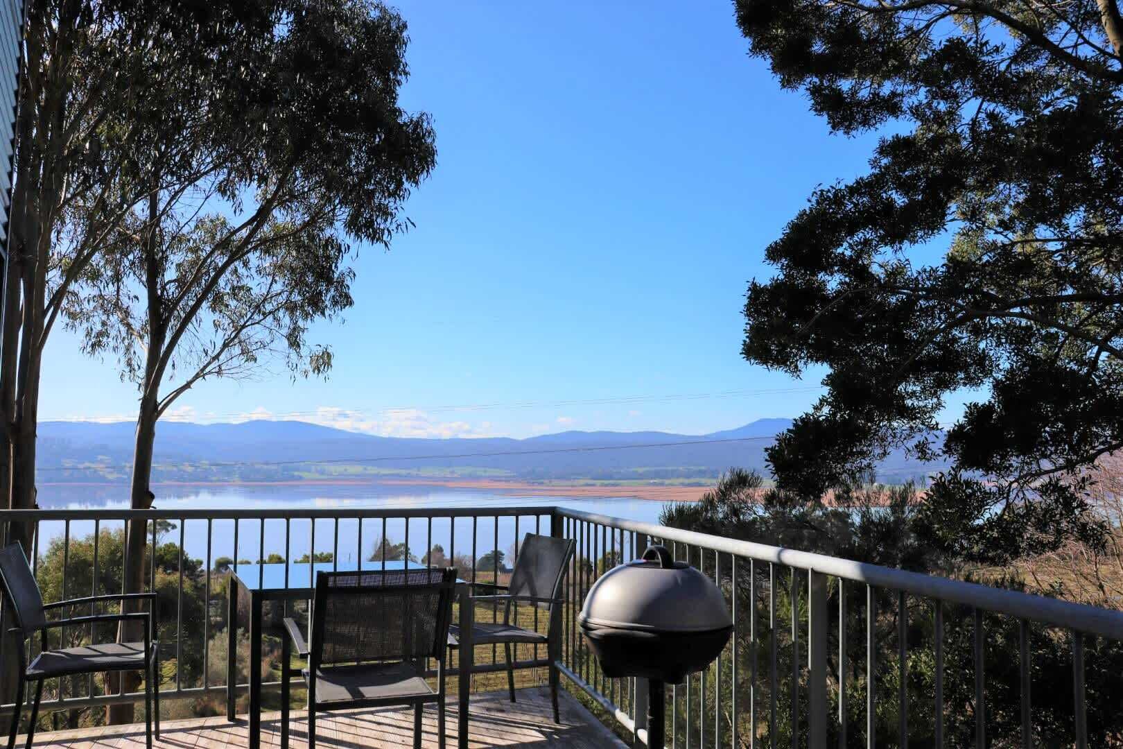 A balcony with chairs and a grill, overlooking a lake and distant mountains.