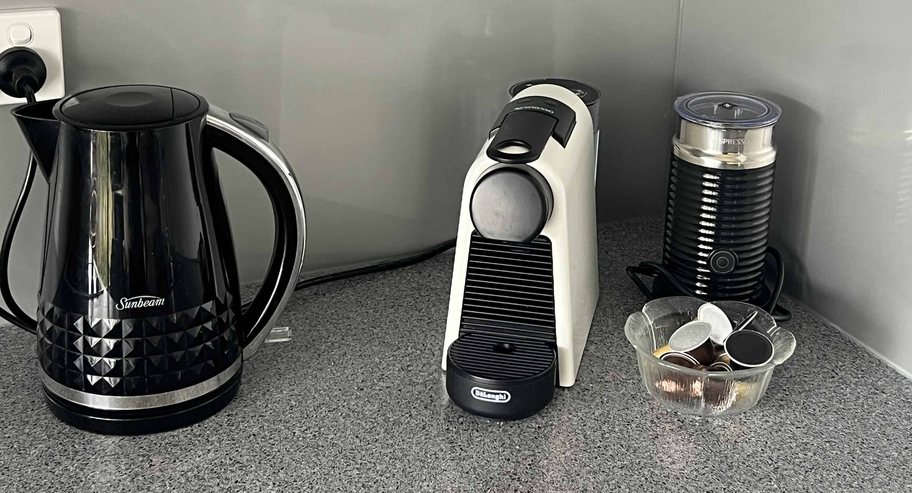A kitchen counter with a black Sunbeam coffee maker, a white and black electric kettle, and a black and silver thermos.