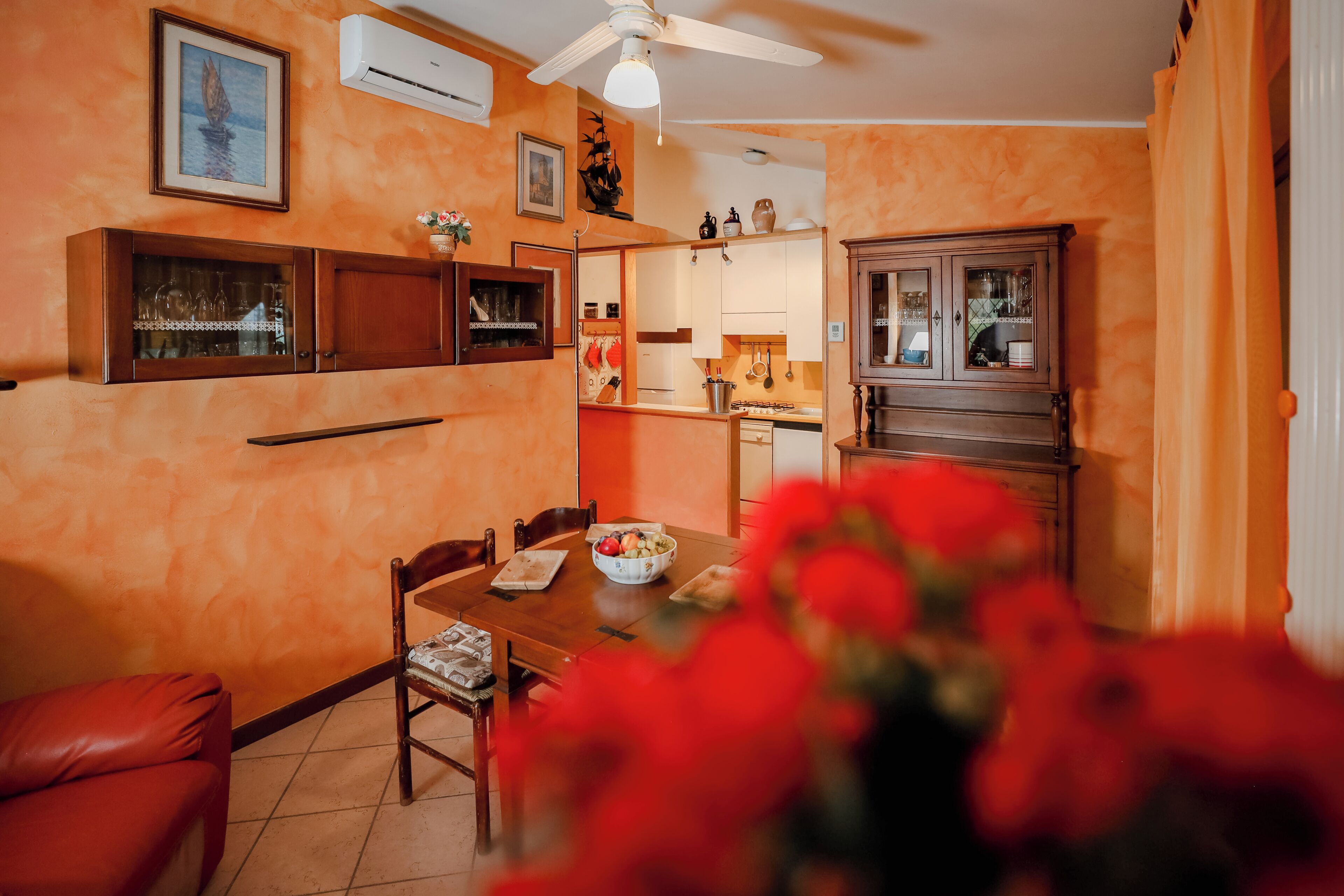 A dining area with a wooden table, chairs, and a vase of red flowers in the foreground.