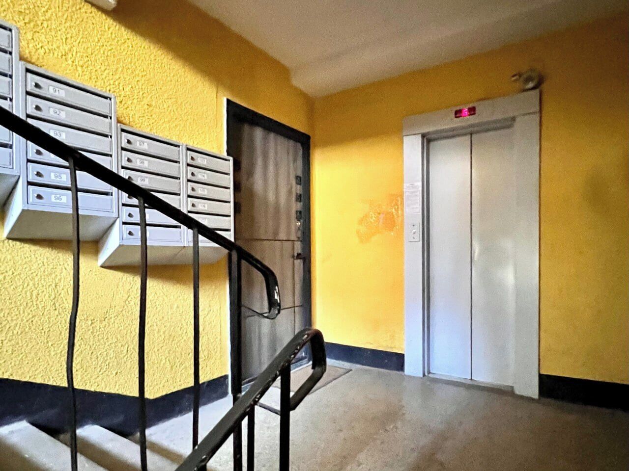 A hallway with a yellow wall, a metal staircase, mailboxes, and an elevator.