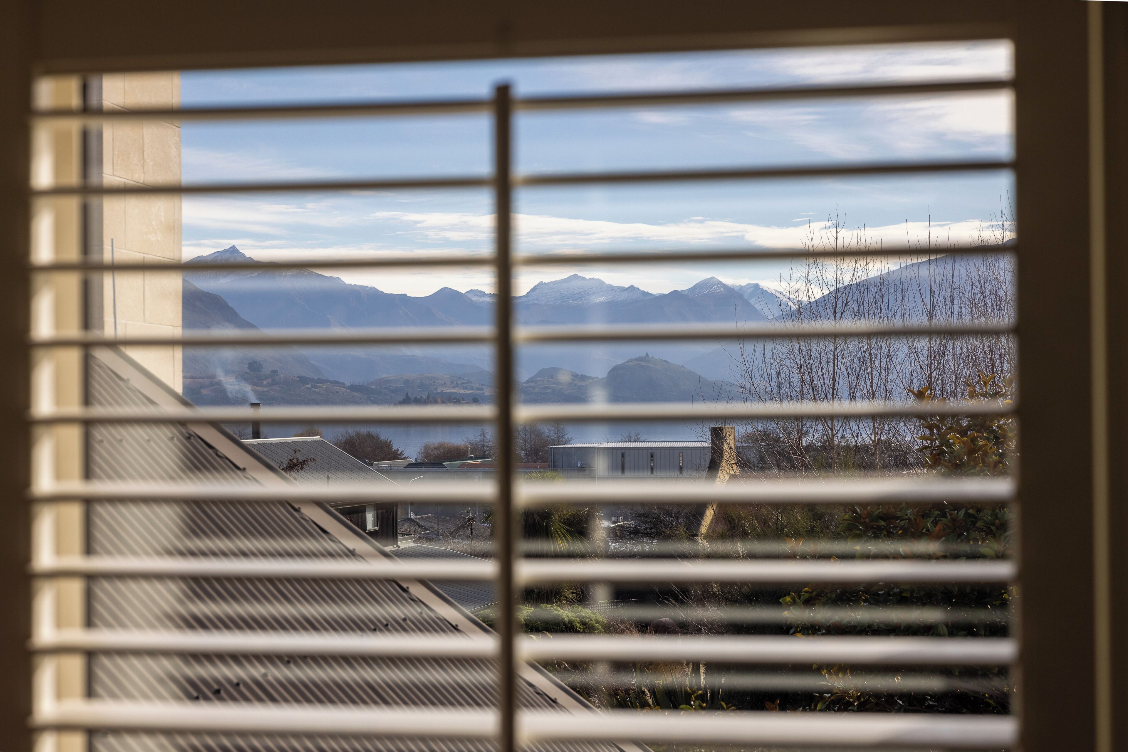 A view through window blinds of a mountainous landscape with buildings in the foreground.