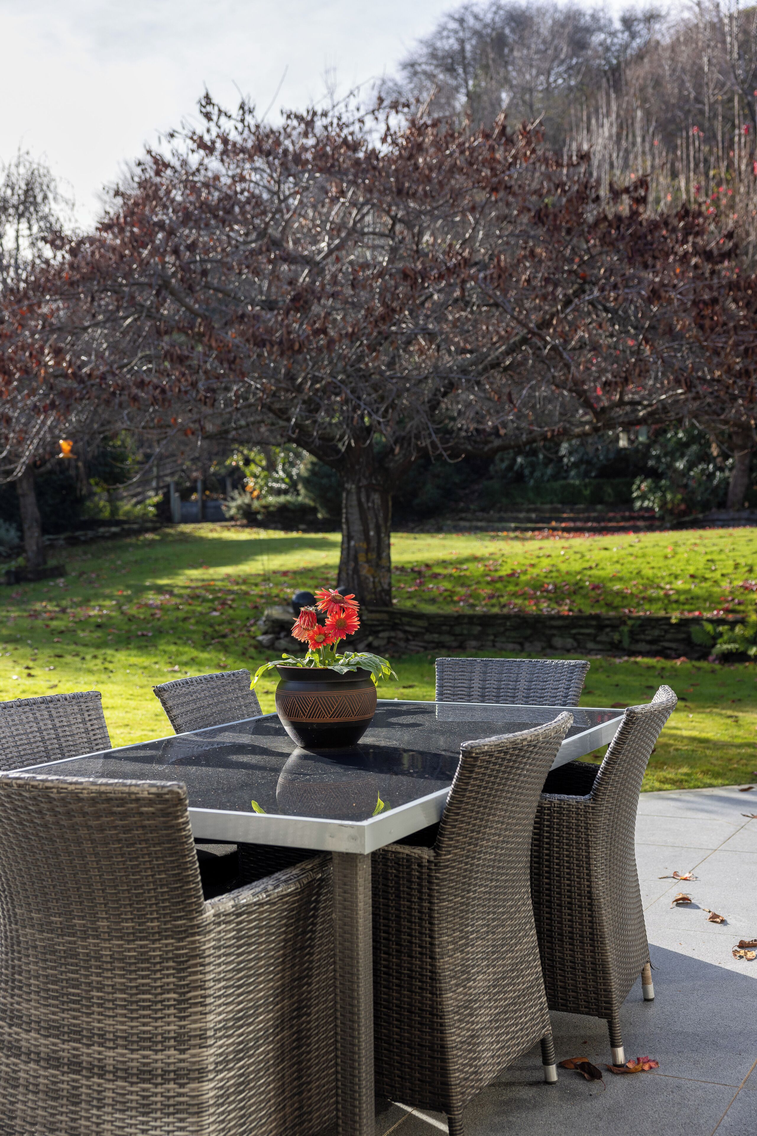 Un área de comedor al aire libre con sillas de mimbre y una mesa de vidrio, rodeada de césped y árboles.