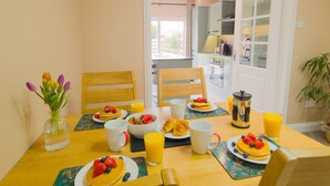 Et morgenbord med pandekager, frugt og juice, en vase med blomster, samt udsigt til et køkken og spiseområde.