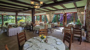 A restaurant with tables set for dining, wooden chairs, and a view of the garden.