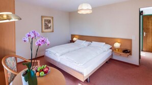 Ein Hotelzimmer mit einem Doppelbett, Nachttischen mit Lampen, einem Sessel, einem runden Tisch mit Obst und einer Topforchidee sowie einem gerahmten Bild an der Wand.