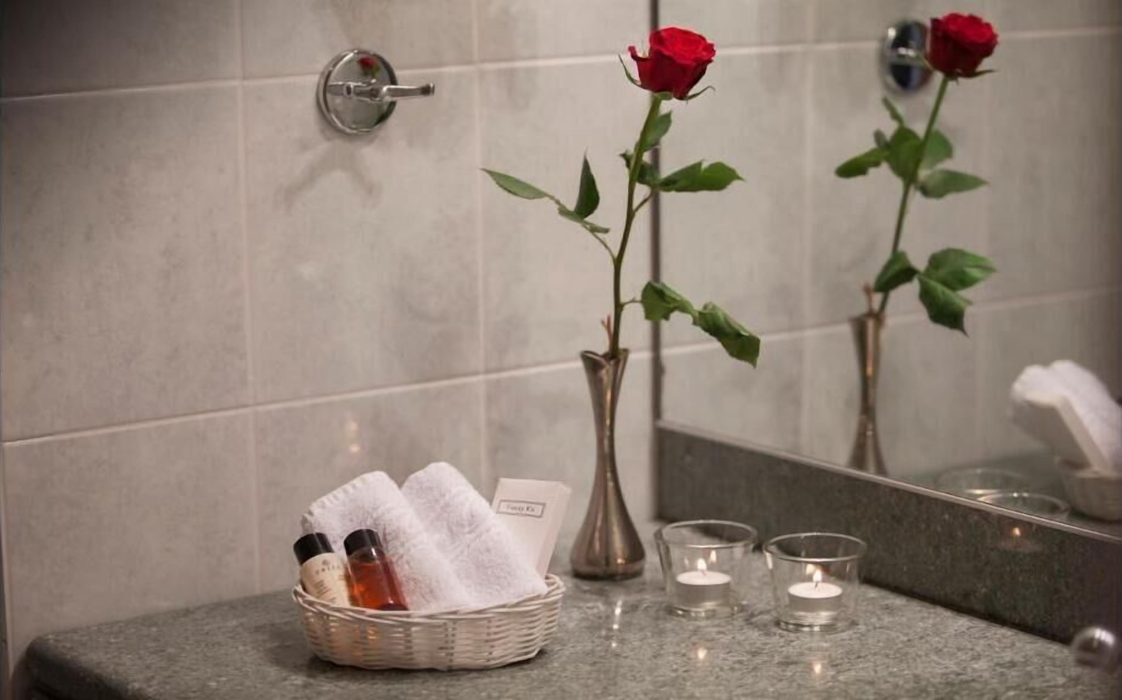 A bathroom with a mirror, a vase with a rose, a basket with towels, and candles.