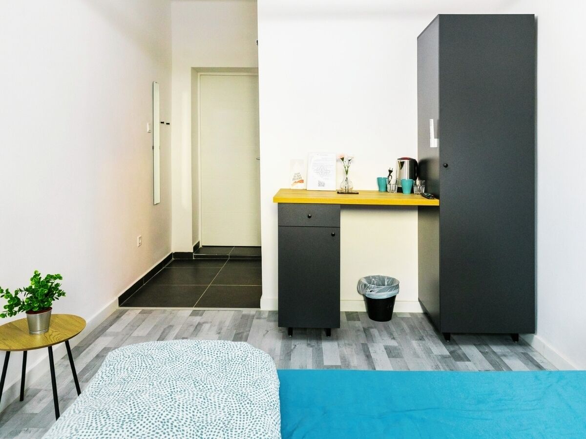 A compact room with a bed, a small desk, a cabinet, and a trash bin.