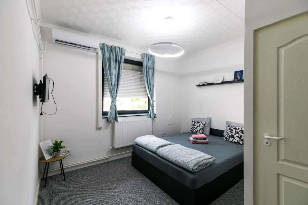A bedroom with a bed, a small table with a plant, a window with curtains, and a shelf with decorative items.
